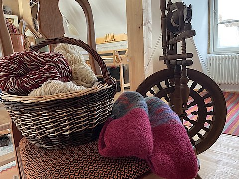 Stuhl mit Korb voll Strickwolle, daneben gestrickte Socken und ein Spinnrad.