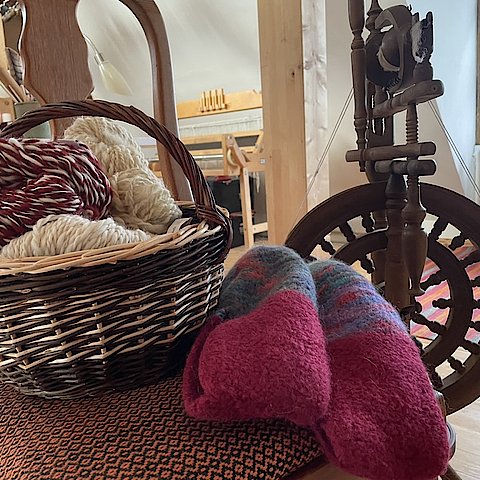Stuhl mit Korb voll Strickwolle, daneben gestrickte Socken und ein Spinnrad.