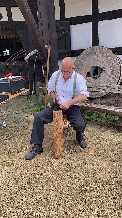 Foto vom Sensen-Workshop-Leiter Uwe K&ouml;rner im sitzen, ein Sensenblatt mit einem Hammer reparierend.